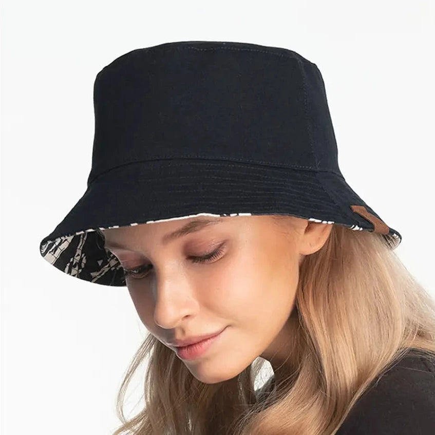 A black bucket hat with a patterned reversible brim, shown on a model. The image features a white border with the word 'REVERSIBLE' printed at the top.