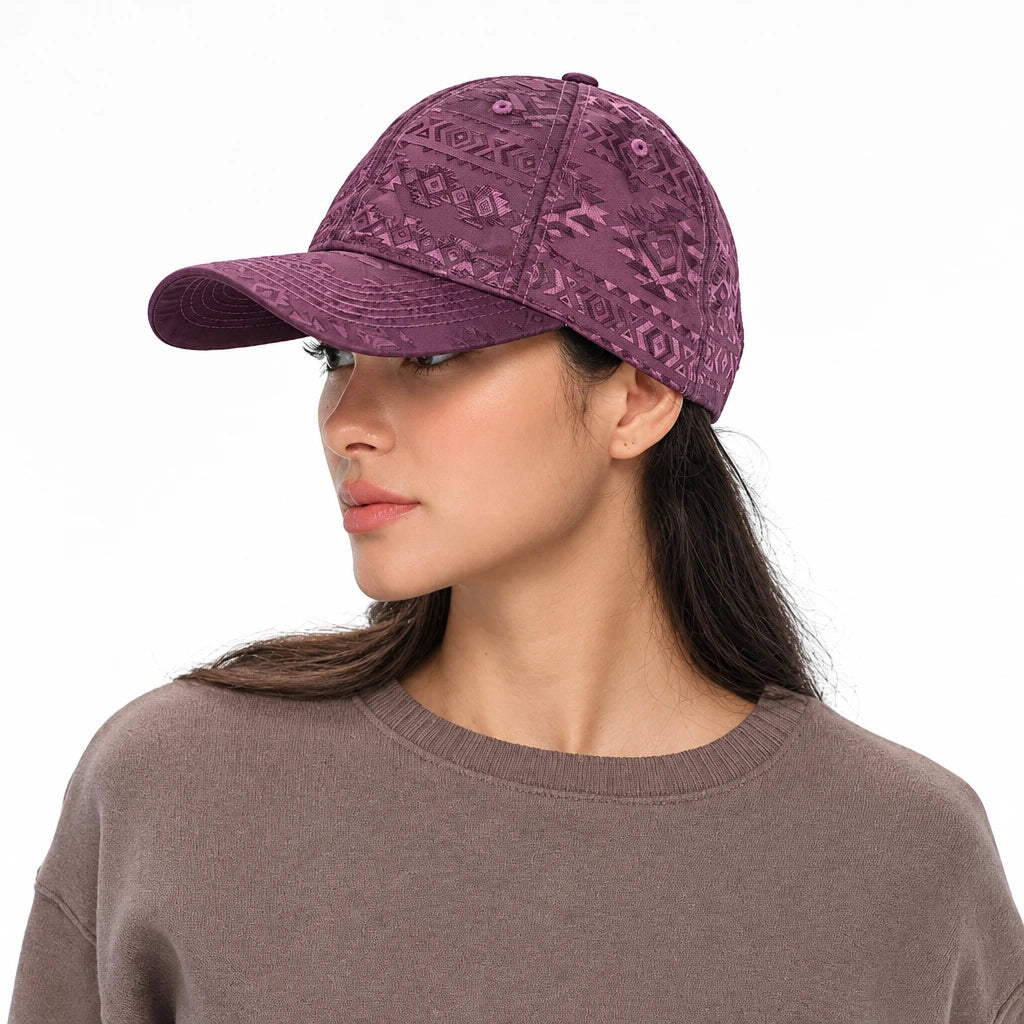 A woman wearing a dusty purple baseball cap with a subtle tonal floral pattern, paired with a brown knit crewneck sweater, set against a plain white background.