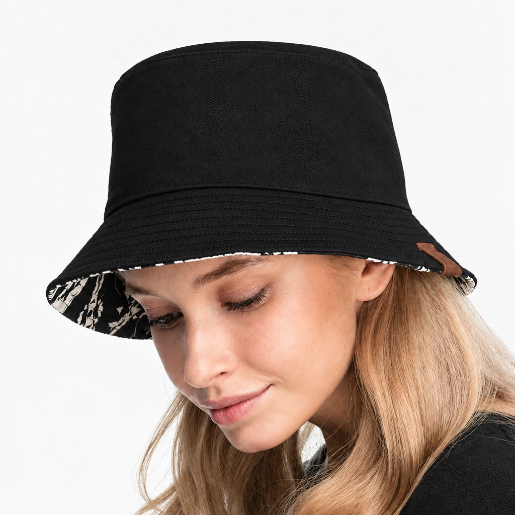 A black bucket hat with a patterned reversible brim, shown on a model. The image features a white border with the word 'REVERSIBLE' printed at the top.