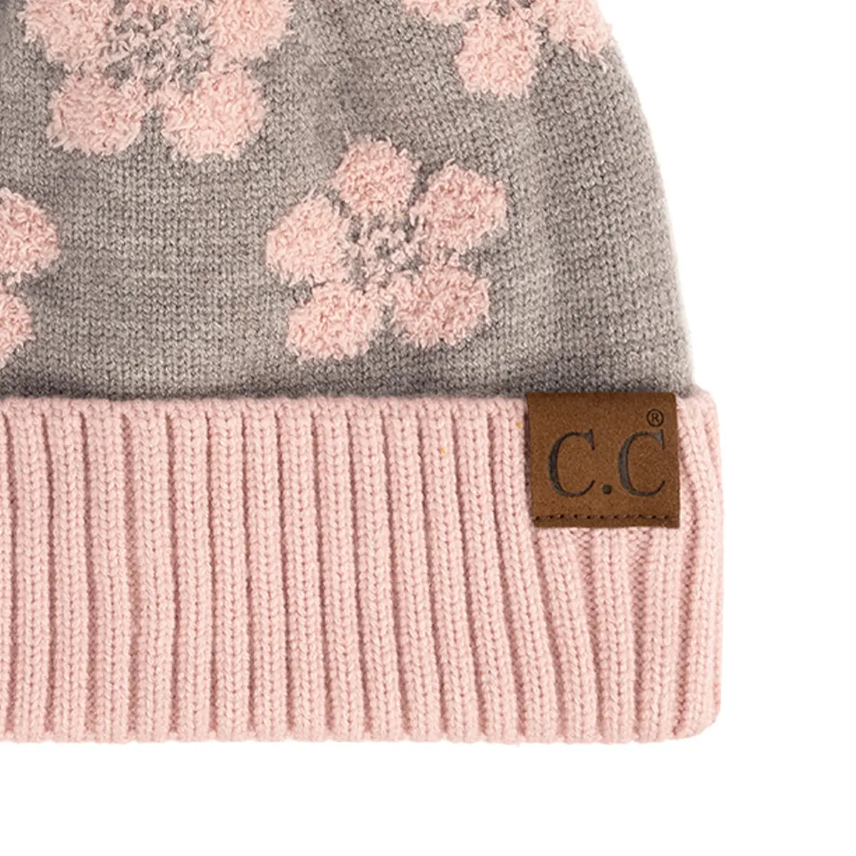Close-up of a knit beanie with a gray upper section featuring pink floral patterns and a light pink ribbed cuff. A small brown leather patch on the cuff is embossed with the letters 'CC'.