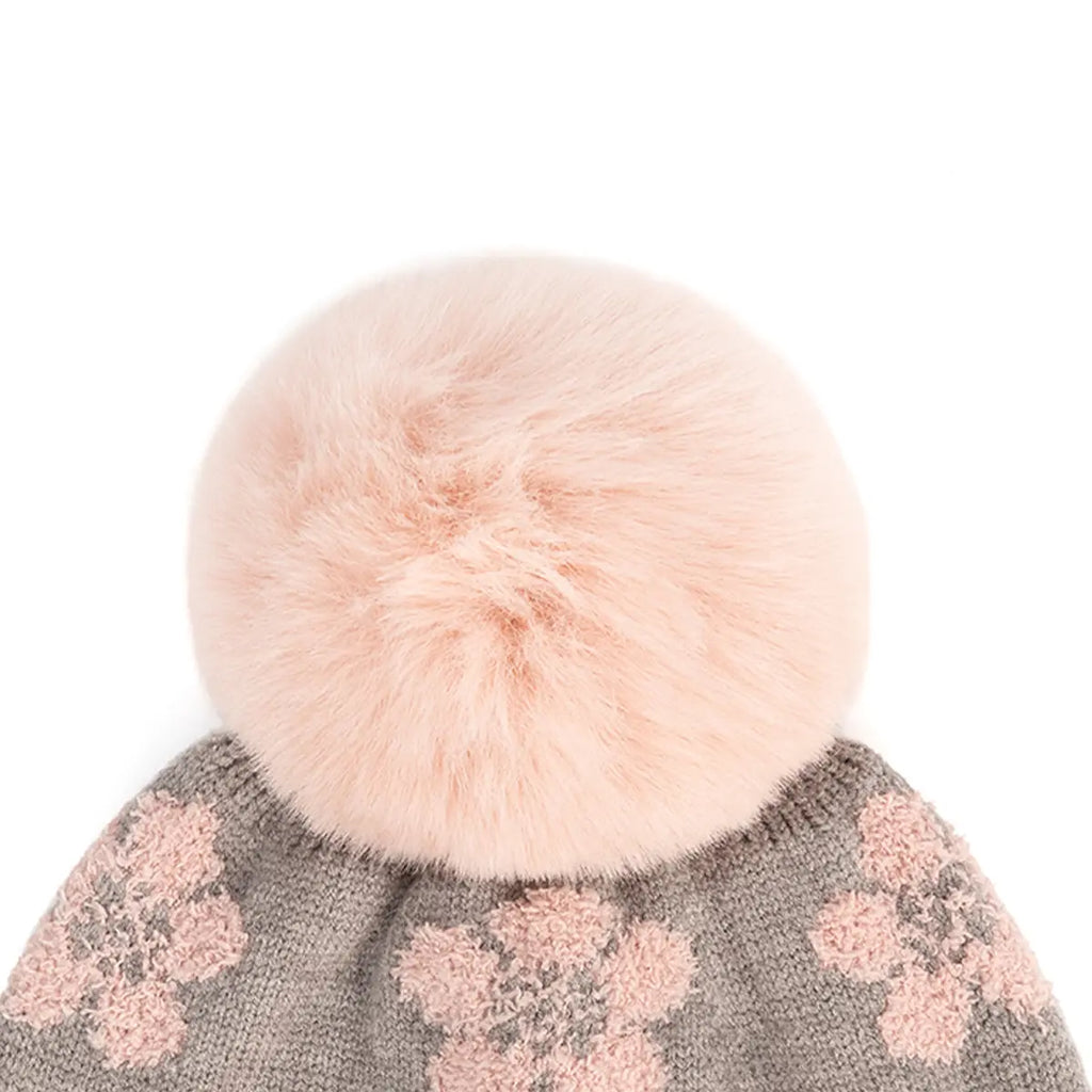 A close-up of a knit winter hat with a large fluffy pale pink faux-fur pom-pom and pink floral knit patterning on a gray background.