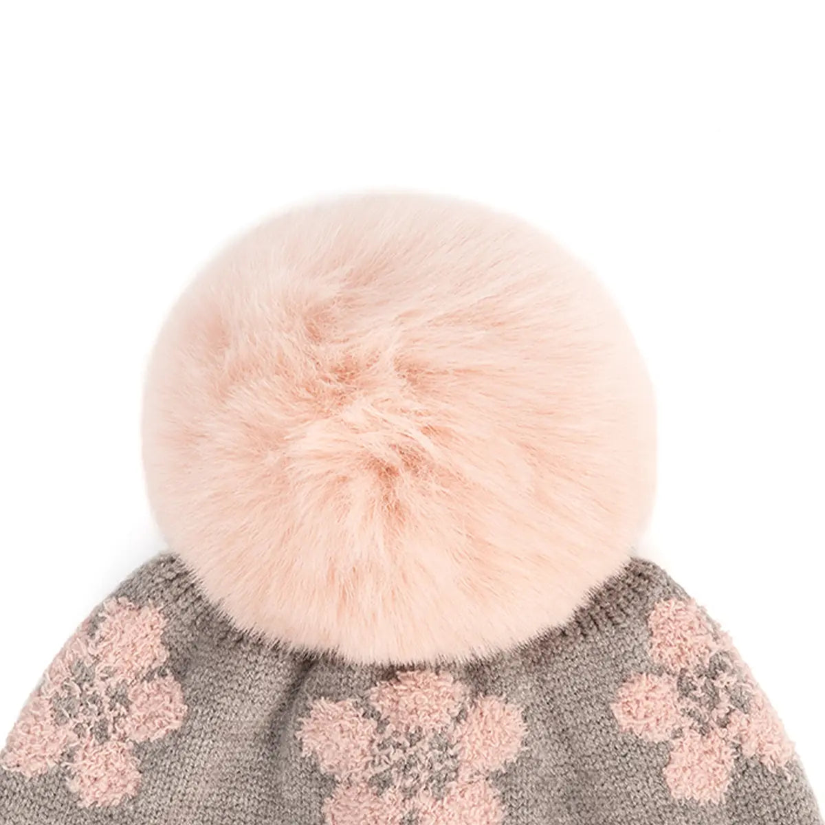 A close-up of a knit winter hat with a large fluffy pale pink faux-fur pom-pom and pink floral knit patterning on a gray background.