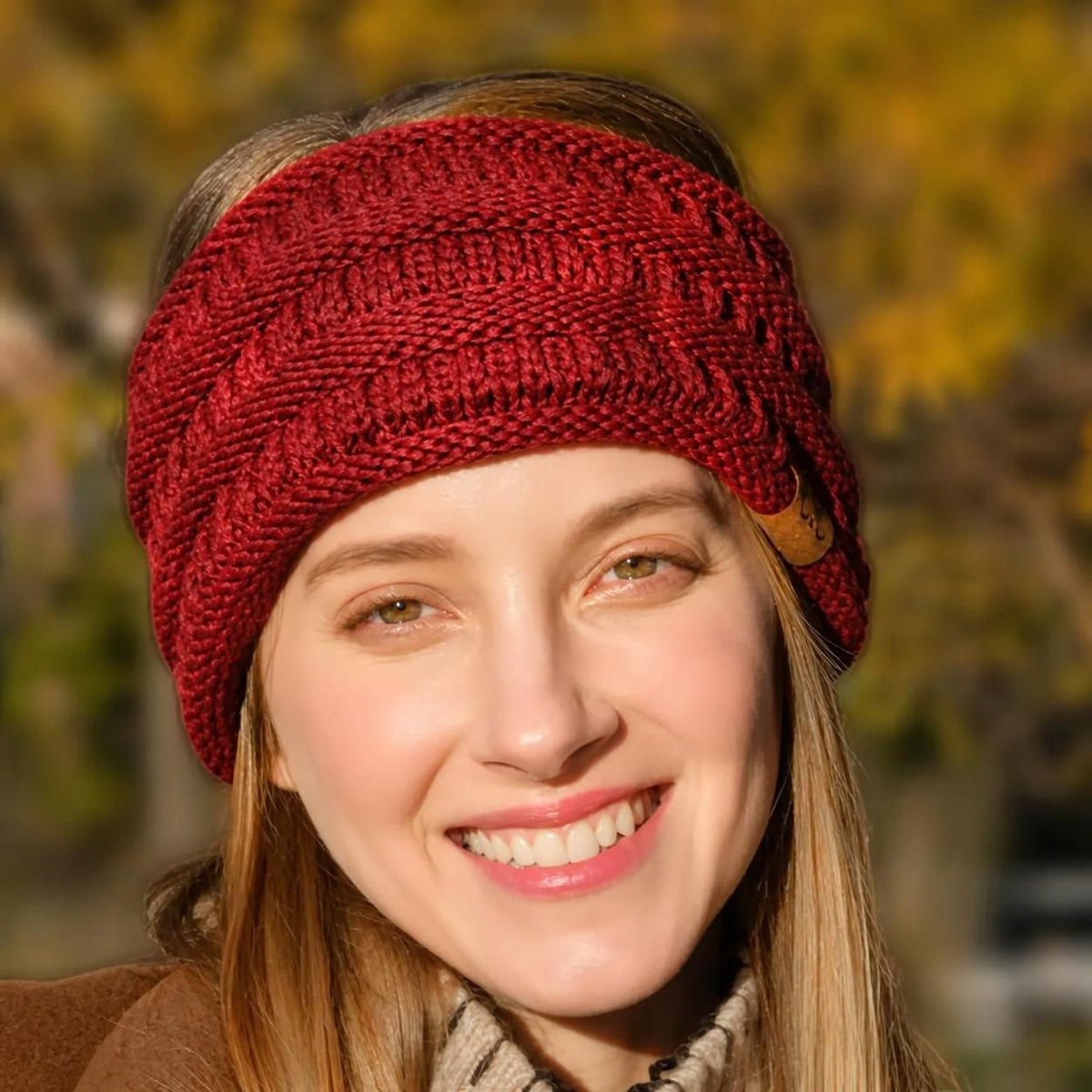 A smiling woman wearing a chunky knit red headband outdoors with a blurred autumn background of yellow trees.