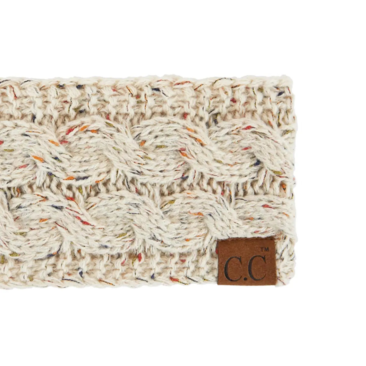 A close-up of a chunky cable-knit headband in cream-colored yarn with subtle multicolor flecks, featuring a brown leather brand patch that reads 'C.C'.
