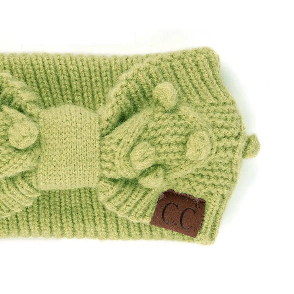 A close-up of a light green chunky knit headband featuring a large decorative bow on top and a small brown leather patch with the letters 'C.C' on the right side, set against a plain white background.
