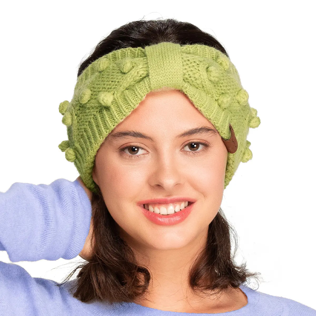 A woman wearing a light green textured knit headband with a central twist detail and pom-pom accents, paired with a light blue sweater against a white background.