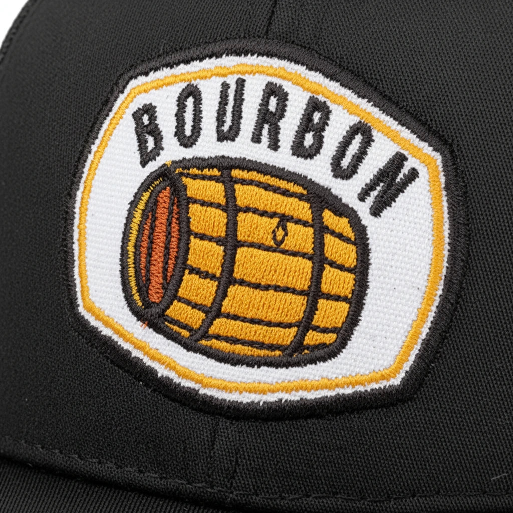 Close-up of a black baseball cap featuring a circular embroidered patch with the word 'BOURBON' in black letters above a yellow barrel graphic on a white background.