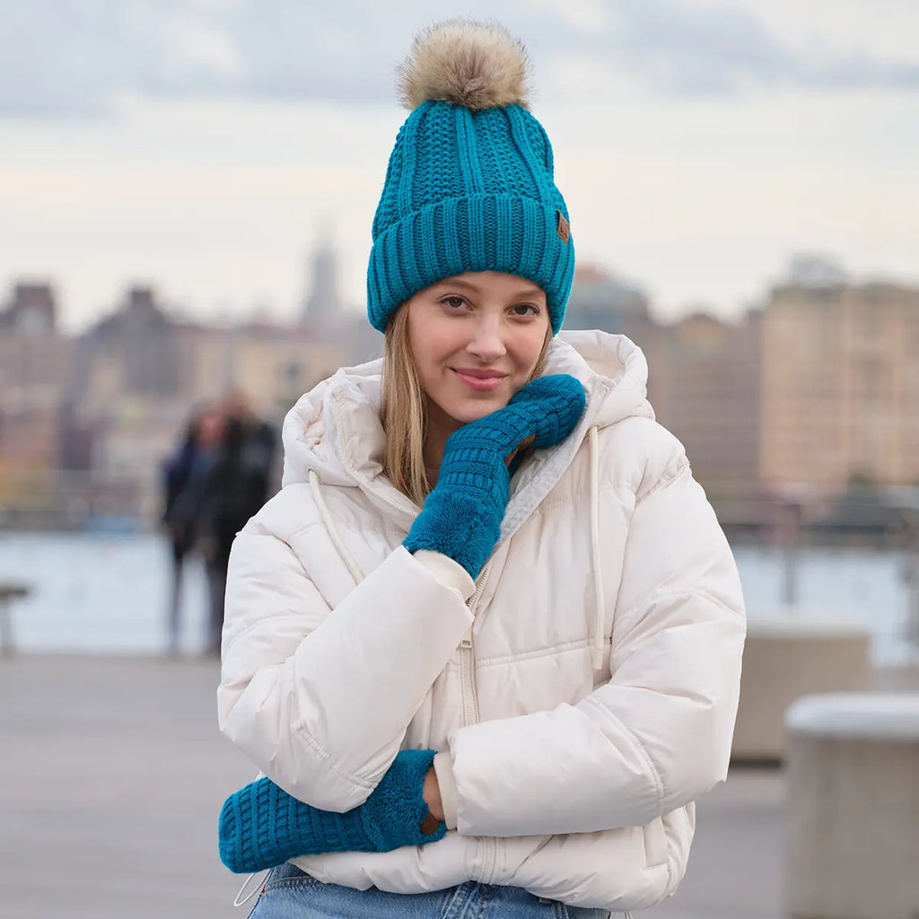 A woman wearing a bright teal ribbed knit beanie with a beige pom-pom, matching teal knit mittens, and a white puffer jacket, standing outdoors with a blurred city waterfront background.