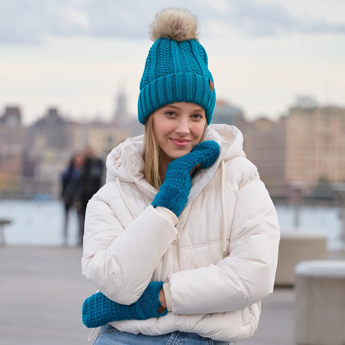 A woman wearing a bright teal ribbed knit beanie with a beige pom-pom, matching teal knit mittens, and a white puffer jacket, standing outdoors with a blurred city waterfront background.