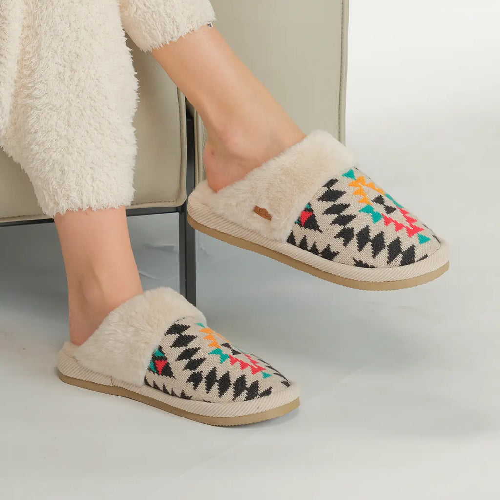 A person wearing fluffy cream-colored slip-on slippers with a colorful geometric pattern on the upper, seated on a light-colored chair.